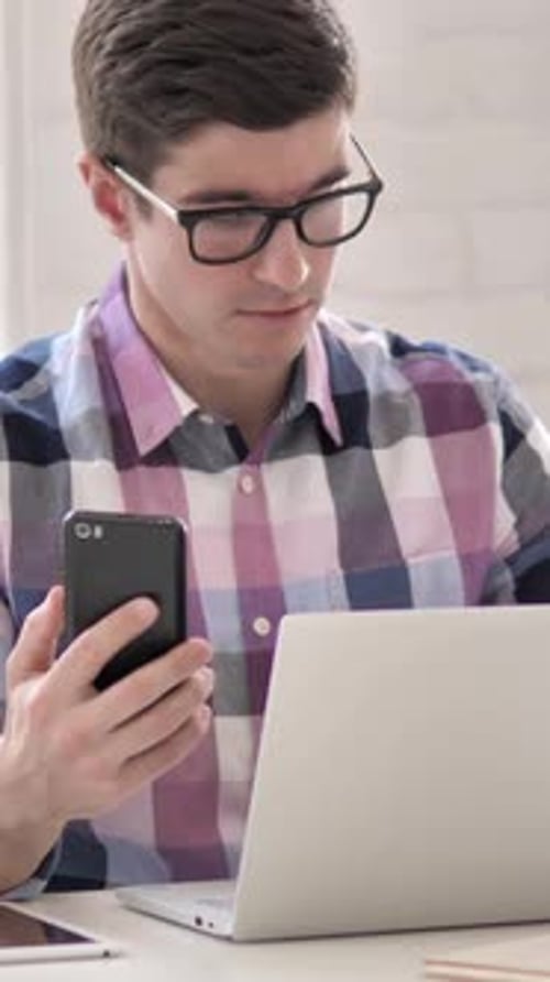 Man in Glasses Using Smartphone with Laptop