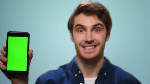 Smiling young man shows mobile phone green screen against blue studio backdrop