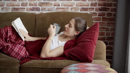 Woman Relaxes on Couch Reading Book and Drinking