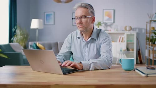 Bonito homem de meia idade usa laptop enquanto está sentado em sua mesa na aconchegante e elegante casa