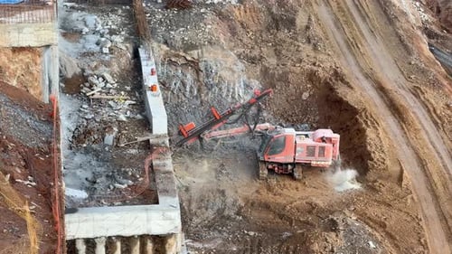 A large, tracked orange drilling rig is boring into a rock surface on a hillside or roadside for