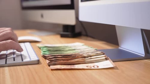 Long row of euro banknotes is spread across a wooden desk in front of computer monitors while someon