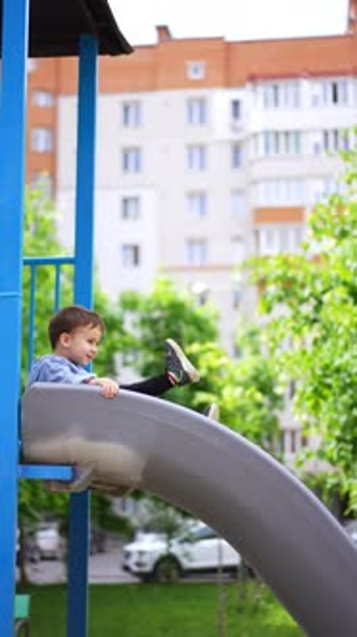 Cheerful smiling baby boy sits on the top of a slide. Kid slides down on the playground.