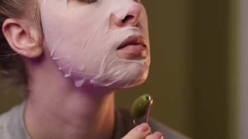 A girl in a fabric face mask massages her skin with a jade roller