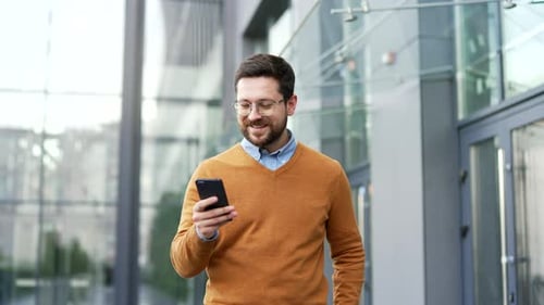 Smiling businessman walking on street holding smartphone outside modern office building. Confident