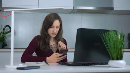Businesswoman Working Remotely From Home Office Using Laptop and Smartphone While Seated at Kitchen