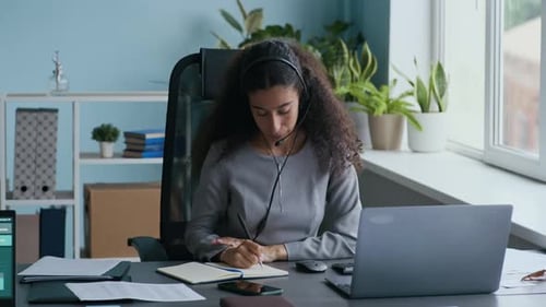 Young Multiethnic Businesswoman Speaking in Headset Microphone and Taking Notes