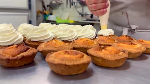 Un pâtissier dans sa boulangerie ajoute de la crème à l'aide d'une poche à douille à des gâteaux récemment préparés sur un plateau en métal