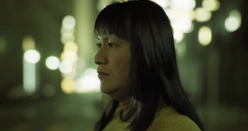 Une personne aux cheveux longs se tient debout tranquillement dans une zone urbaine faiblement éclairée
