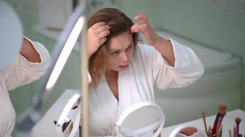 A Woman in a White Bathrobe Combs the Hair on Her Head Near Her Forehead with Her Finger Sitting in