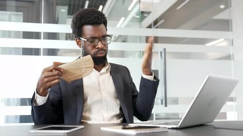 African american businessman in formal suit experiencing discomfort at work uses fan to cool down