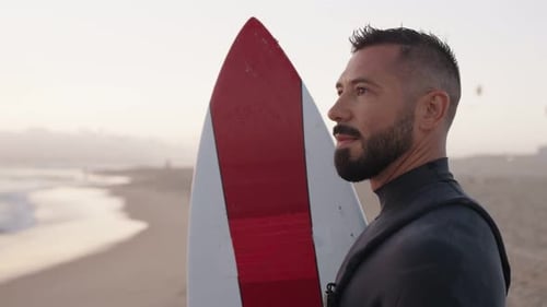 Surfista contemplativo em roupa de mergulho segura prancha de surfe na praia ao pôr do sol
