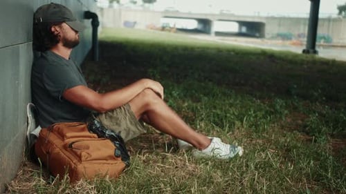 Tired Homeless Tourist Sleeps on a Bench in the Park Putting a Backpack Under His Head