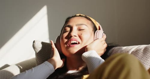 Mid-adult Asian woman lifting pink headphones and adjusting mustard headband on cushion humming