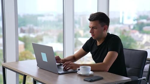 Junger konzentrierter Mann, der im Büro am Laptop arbeitet. Mann trinkt eine Tasse Kaffee, schaut auf den Bildschirm