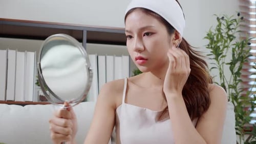 Young asian woman looking mirror with stress on facial sitting on sofa in living room at home.