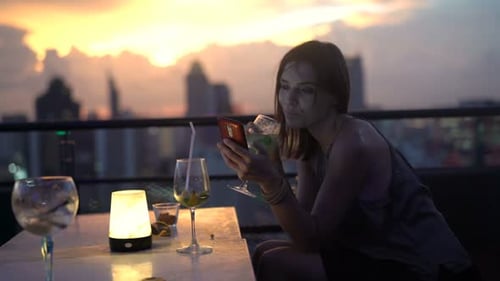 Young Woman Texting on Smartphone and Drinking Cocktail in Skybar During Sunset Adult