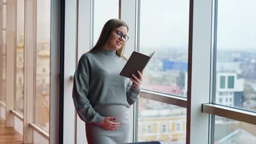 Positive smiling pregnant woman wearing glasses reads a book.