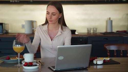 Young Woman Drinks Orange Juice and Works on Her Laptop at Breakfast at Home