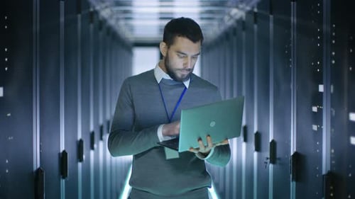 Male IT Engineer Works on a Laptop in a Big Data Center. Rows of Rack Servers are Seen.