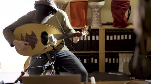 With computer in foreground, young man playing oud records music with doumbek drums in background as