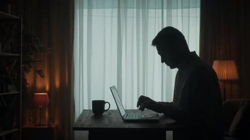 Side View of a Dark Silhouette of a Man at a Table in Front of a Laptop A Man Works on a Laptop with