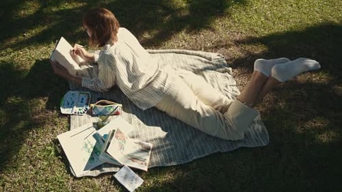 An Inspired Woman Lying on the Blanket in the Blooming Park and Drawing in Her Sketchbook