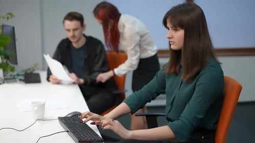 Young Caucasian Focused Woman Typing on Computer Keyboard As Blurred Colleagues Talking at