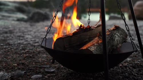 Big wooden fire logs burning on stone gravel, camp fire in front of lake