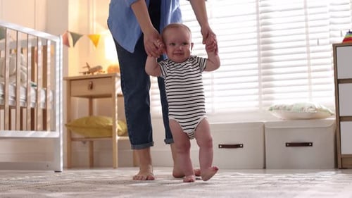 First steps. Mother supporting daughter while she learning to walk at home, closeup