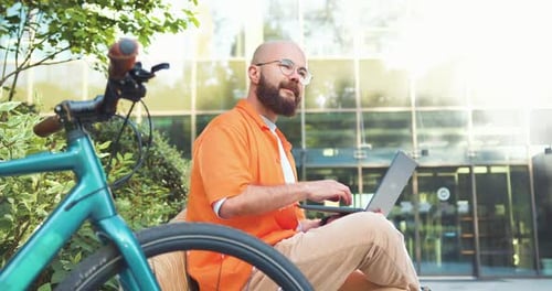 Elegante homem hipster de óculos trabalhando no laptop enquanto está sentado na praça da cidade no banco com bicicleta