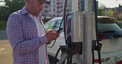 Man with Smartphone Waits While His Electric Car Charging at a Charging Station