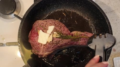 Cooking of a big red beef stake in a black pan with onion and rosemary