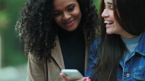 Two women laughing together while using phone outside