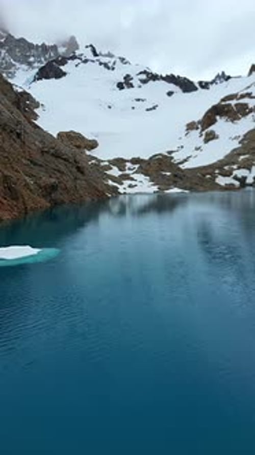 Aerial drone view of a turquoise mountain lake surrounded by rugged terrain and snowfields beneath t