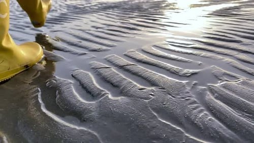 Sun reflection on a rippled sandy beach and yellow boots walking in slow motion