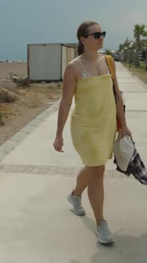 Vertical video. A Woman Walks Along a Paved Path Near Tennis Courts, Wrapped in a Yellow Towel After