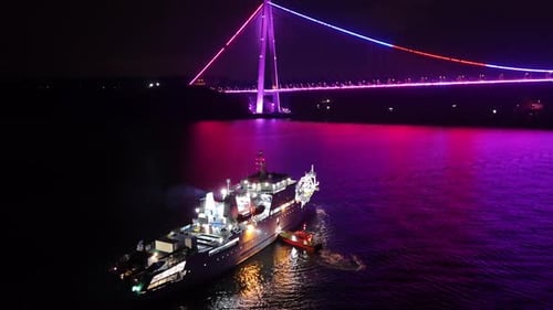 Ship and Tugboat at Night Under Lit Bridge