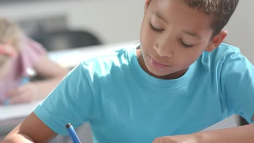 Ein biracialer Junge in einem hellblauen Hemd konzentriert sich auf das Schreiben in einem Klassenzimmer der Schule