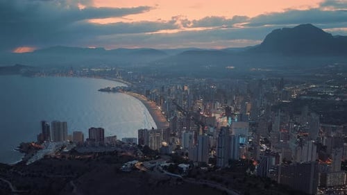 Modern coastal city next to the blue ocean and mountains with orange sunset in the background.