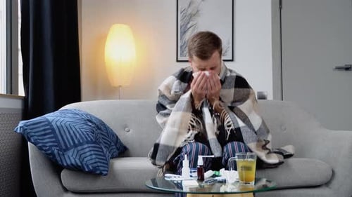 Lustiger fröhlicher junger Mann sitzt zu Hause auf der Couch unter einem Plaid Niesen in einer Serviette fühlt sich schwach