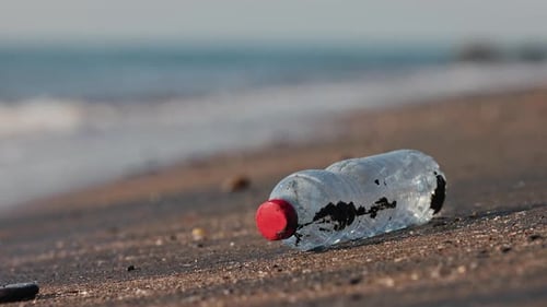 Plastikflasche an einem Sandstrand, der in Zeitlupe von Meereswellen umspült wird, Umweltverschmutzungskonzept