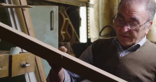 Senior Man Inspects Piece of Wood in Workshop