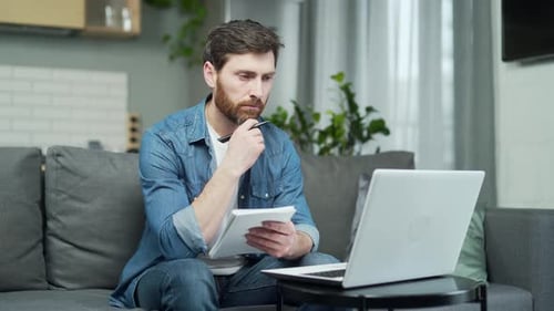 Concentrated caucasian man writing down in the notebook from laptop computer. Male freelancer