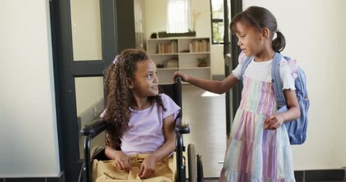 Diverse female child friends meeting after school by wheelchair with blue pack touching it smiling