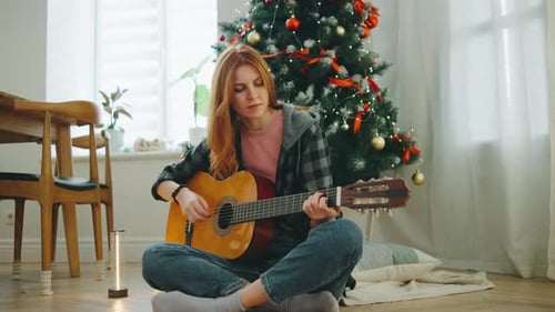 Young Woman Playing Guitar While Sitting on the Floor Next to a Decorated Christmas Tree in a Cozy