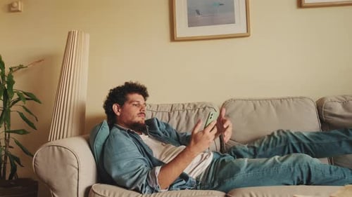 Junger Mann tippt Textnachricht, während er auf dem Sofa liegt