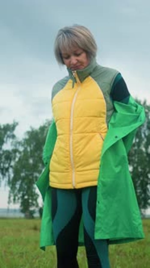 Woman Removes Green Raincoat in Open Grassy Field Under Cloudy Sky