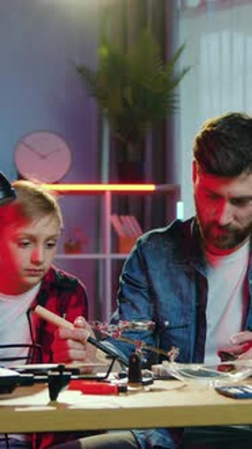 Handsome Attentive Teen Boy Sitting at the Table Together with His Goodlooking Skillful Adult