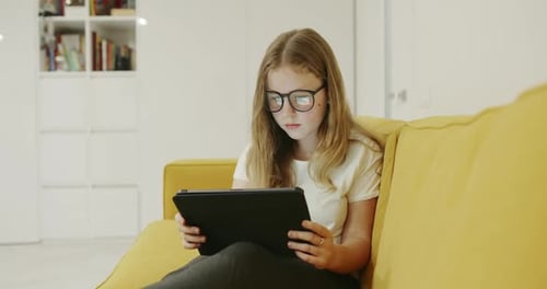 Young Blond Girl Wearing Glasses Sitting on a Comfortable Sofa at Home Using a Tabletpc During the